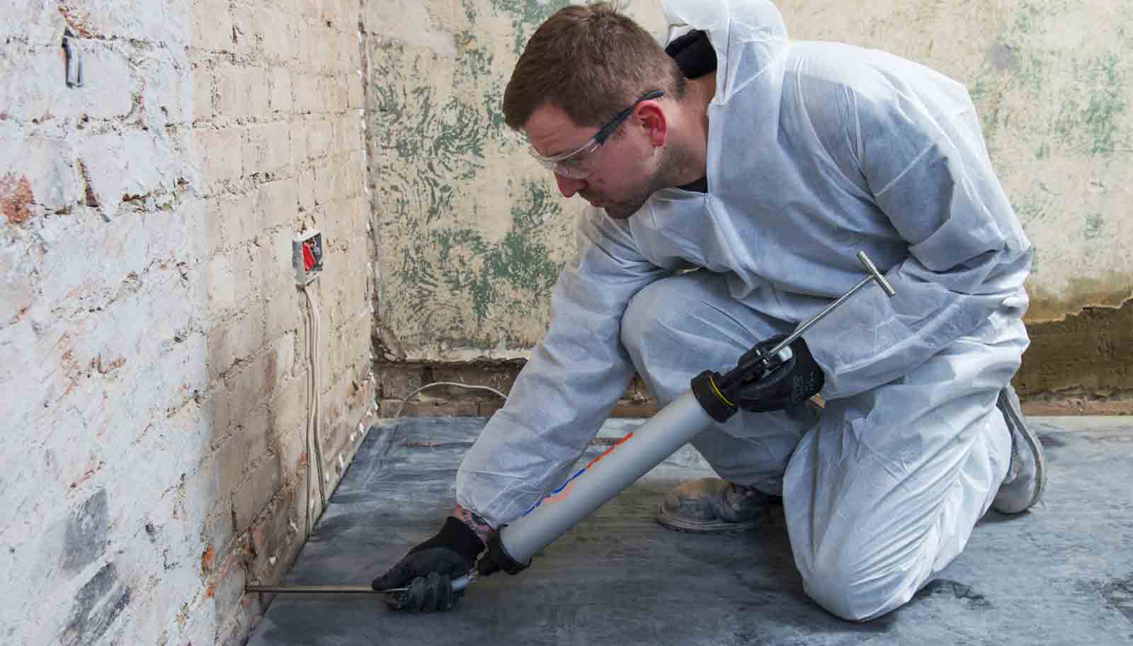 Un expert en humidité, tout en combinaison de protection, est agenouillé pour injecter un produit dans un mur en briques.